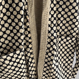 Peut inclure: Longue veste ouverte &agrave; l'avant, orn&eacute;e de pois noirs et blancs. La veste comporte deux poches avant et une doublure cr&egrave;me. Le tissu semble &ecirc;tre matelass&eacute; avec des coutures visibles.