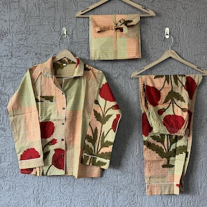 May include: A long-sleeve button-down shirt and matching pants set featuring a yellow and orange checkered pattern with large red floral prints and green leaves. A folded fabric piece with the same pattern is displayed on a wooden hanger.