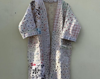 Puede incluir: Bata larga con diseño patchwork y abertura frontal. La bata es principalmente blanca con varios estampados florales y geométricos en rojo, azul, verde y negro. Tiene bolsillos y está colgada de una percha de madera.
