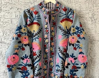 Può includere: Giacca ricamata azzurra con collo alto. La giacca presenta ricami floreali colorati in rosa, giallo, rosso e blu. La giacca è appesa a una gruccia bianca contro un muro strutturato.