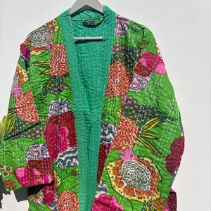 Peut inclure: Une robe matelassée verte ornée d'un motif floral aux tons rose, orange et jaune. La robe est doublée et bordée de vert contrastant. Le vêtement est suspendu à un cintre gris.