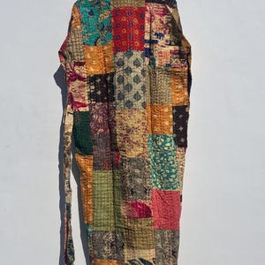 May include: A patchwork robe with a variety of colorful fabric squares. The robe is hanging on a wooden hanger against a white wall. The robe has a long tie belt and is made of various patterned fabrics in shades of red, green, orange, and brown.