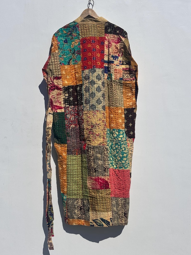 May include: A patchwork robe with a variety of colorful fabric squares. The robe is hanging on a wooden hanger against a white wall. The robe has a long tie belt and is made of various patterned fabrics in shades of red, green, orange, and brown.