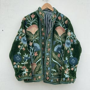Peut inclure: Veste en velours vert foncé avec broderies florales pêche, bleues et vertes. La veste a un col montant et une doublure rayée. La broderie détaillée recouvre le devant, les manches et le col de la veste.
