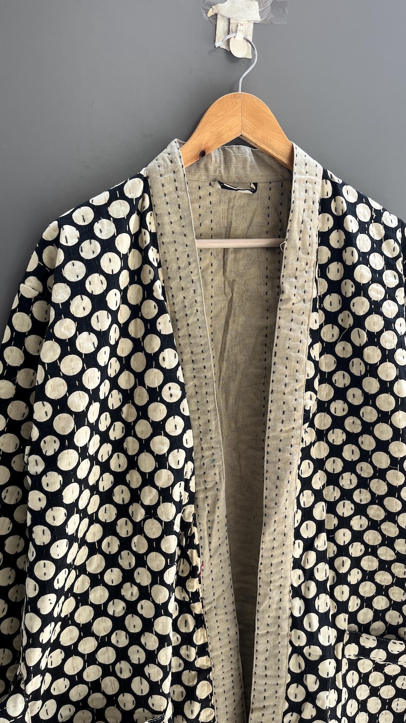 Peut inclure: Une veste de style kimono &agrave; motifs avec un int&eacute;rieur cr&egrave;me et un ext&eacute;rieur &agrave; pois noirs et cr&egrave;me. La veste est suspendue &agrave; un cintre en bois sur un fond gris. Les bords sont cousus.
