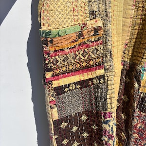 May include: A patchwork jacket in shades of tan, brown, and burgundy. The jacket features a variety of patterned fabrics, including floral and geometric designs, stitched together with visible thread. The jacket has a pocket on the front.