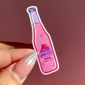 May include: A pink sticker in the shape of a bottle of Jarritos soda. The sticker has a purple top and a label with mushrooms and the Jarritos logo. Two hearts and two stars are below the logo.
