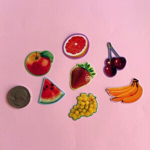 May include: A collection of seven fruit stickers including a red apple, a grapefruit slice, a pair of cherries, a strawberry, a watermelon slice, a bunch of yellow grapes, and a cluster of bananas, shown next to a US quarter coin for scale.