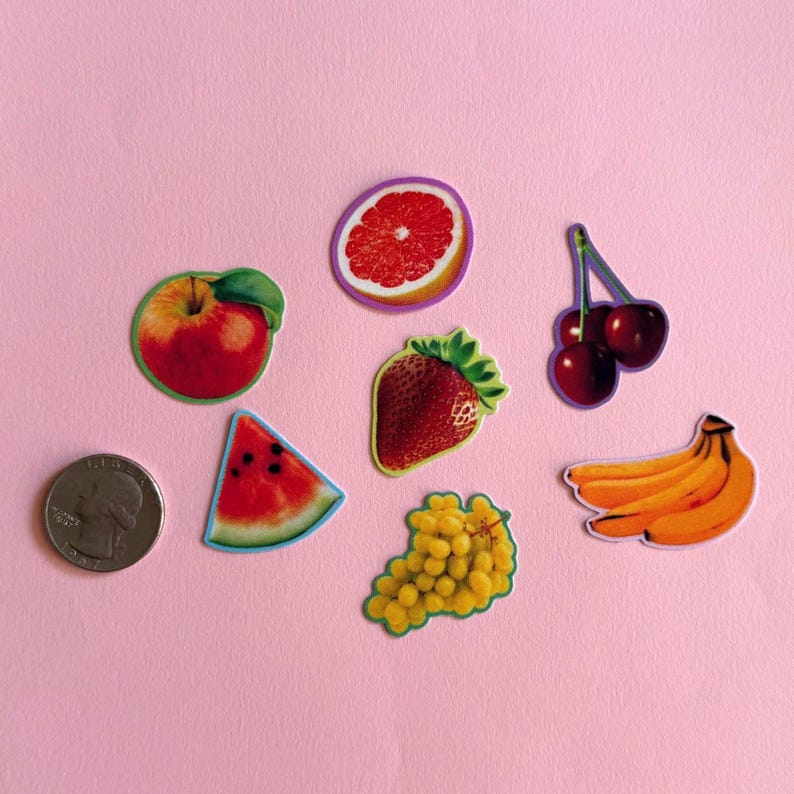 May include: A collection of seven fruit stickers including a red apple, a grapefruit slice, a pair of cherries, a strawberry, a watermelon slice, a bunch of yellow grapes, and a cluster of bananas, shown next to a US quarter coin for scale.