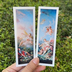 May include: Two rectangular bookmarks featuring classical rococo-style paintings. The left bookmark depicts figures in a landscape with blue skies and clouds. The right bookmark shows cherubs and a central figure amidst a sky and water scene.