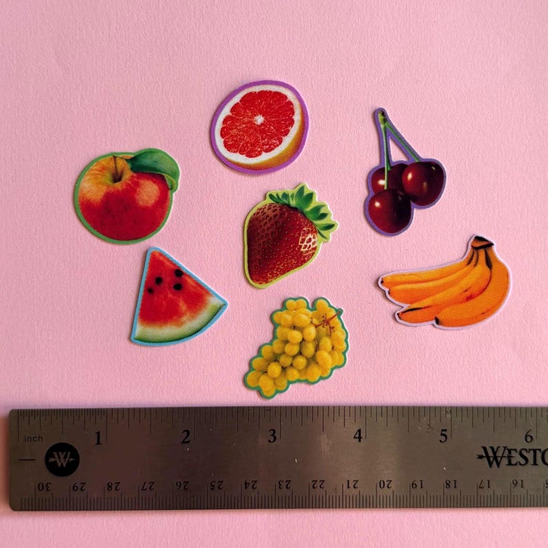 May include: A collection of fruit stickers including a red apple, grapefruit slice, strawberry, cherries, watermelon slice, yellow grapes, and bananas, positioned above a metal ruler measuring approximately 6 inches in length.