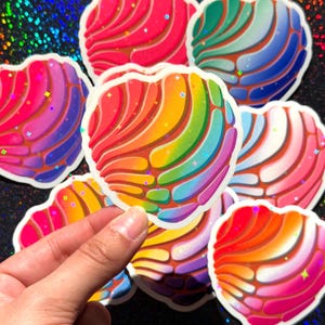 May include: A collection of heart-shaped stickers featuring a concha bread pattern with a vibrant rainbow gradient. Each sticker has a glossy finish with holographic star accents scattered across the surface, showcasing various color combinations.