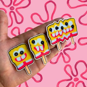 May include: Three yellow, red, and white cartoon character popsicle stickers on a pink background. Each sticker features a different design of the character with black eyes and a red smile. The popsicles are on wooden sticks.