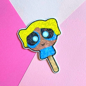 May include: A glittery sticker in the shape of a cartoon character popsicle. The character has yellow hair, blue eyes, and a blue dress. The popsicle stick is light brown, and the background is pink and white.