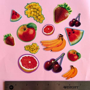 May include: A collection of fruit stickers including grapes, strawberries, cherries, apples, bananas, watermelon slices, and grapefruit halves, arranged above a metal ruler showing measurements in inches for scale and size reference.