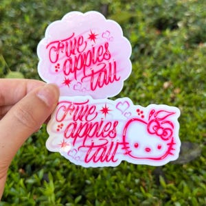 May include: Two die-cut vinyl stickers with a light pink background and bright pink cursive text that reads Five apples tall. The bottom sticker features a white Hello Kitty character outline with a bow, surrounded by small hearts and star accents.