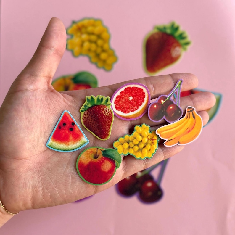 May include: A collection of small fruit-themed stickers featuring a watermelon slice, a strawberry, a grapefruit slice, a bunch of yellow grapes, a bunch of cherries, and a cluster of yellow bananas, all displayed together in a palm.