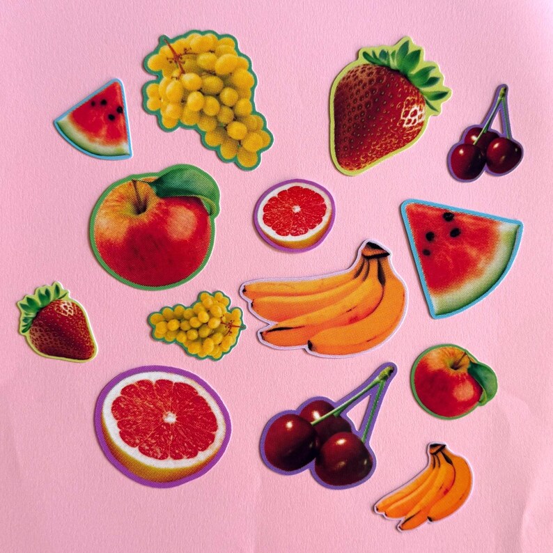 May include: A collection of fruit stickers featuring a watermelon slice, green grapes, a strawberry, cherries, a red apple, a grapefruit slice, a bunch of bananas, a smaller strawberry, a smaller grape cluster, and a smaller apple on a pink background.