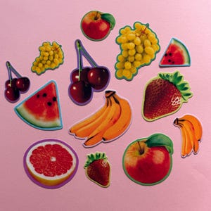 May include: A collection of assorted fruit stickers featuring a red apple, green and red grapes, a watermelon slice, cherries, yellow bananas, a strawberry, a grapefruit slice, and a red apple with a green leaf, all arranged on a pink surface.