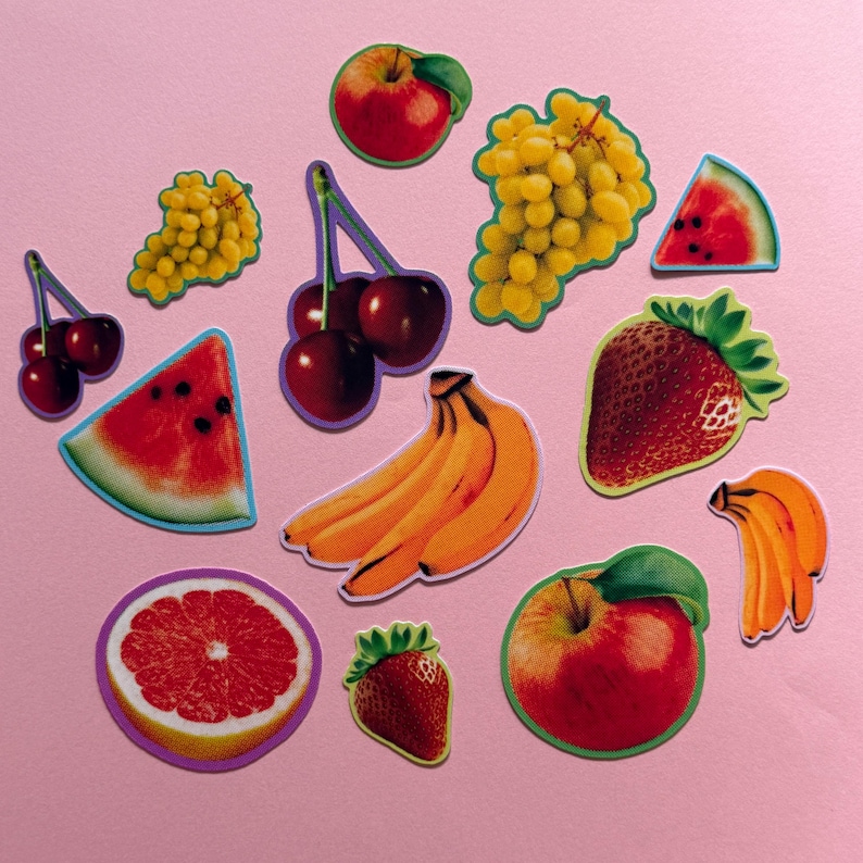 May include: A collection of assorted fruit stickers featuring a red apple, green and red grapes, a watermelon slice, cherries, yellow bananas, a strawberry, a grapefruit slice, and a red apple with a green leaf, all arranged on a pink surface.