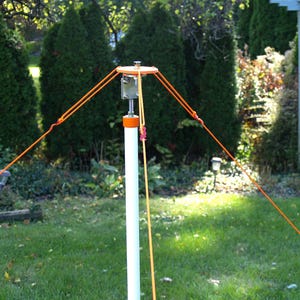 May include: A white pole with orange accents and guy lines, set up outdoors. The pole has a metal top with orange ropes extending outwards. The background features green trees and grass, suggesting a backyard setting.
