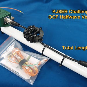 Puede incluir: Antena vertical OCF Halfwave KJ6ER Challenger, longitud total 58 cm. La antena es blanca con detalles en naranja y verde, un cable negro y una caja verde con la etiqueta "LDG". Una bolsa transparente contiene cables.