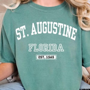 Può includere: Una t-shirt a maniche corte verde chiaro con testo bianco che recita ST. AUGUSTINE FLORIDA e un riquadro rettangolare contenente il testo EST. 1565. La maglietta ha un girocollo e una vestibilità ampia, con la scritta LIGHT GREEN in nero.