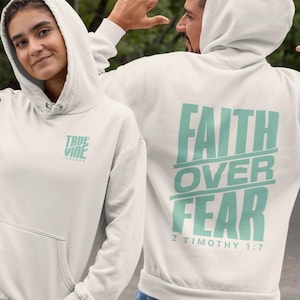 May include: Off-white hooded sweatshirts. The front features the text "TRUE VINE" in light teal, while the back displays "FAITH OVER FEAR" and "2 TIMOTHY 1:7". They have a front pocket and a drawstring hood.