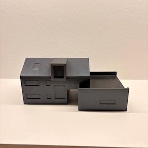 Könnte beinhalten: Graues 3D-gedrucktes Architekturmodell eines modernen Hauses. Das Modell verfügt über eine rechteckige Hauptstruktur mit Flachdach, einen kleinen quadratischen Turm und einen rechteckigen Anbau. Das Haus hat mehrere rechteckige und quadratische Aussparungen.
