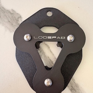 Könnte beinhalten: Schwarzes Metallwerkzeug mit dem Text "LOOSFAB" und drei kreisförmigen Komponenten. Das Werkzeug hat eine einzigartige Form mit einem Loch oben und ist mit silbernen Schrauben befestigt. Der Hintergrund ist eine helle, gemusterte Oberfläche.