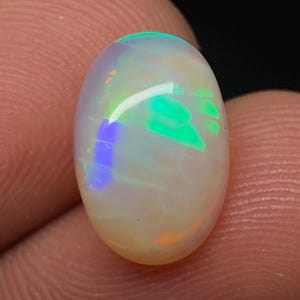 Op de afbeelding: Een ovaalvormige cabochon opaal edelsteen met een doorschijnende witte basis en levendige flitsen van neongroen, elektrisch blauw en subtiel oranje kleurspel over het gladde, gepolijste en gewelfde oppervlak tegen een donkere achtergrond.