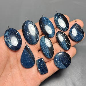 May include: A collection of ten polished gemstones in various shapes, including oval, teardrop, and rectangular. The stones feature a dark blue base with white and light blue swirling patterns and speckled accents, resembling a starry night.