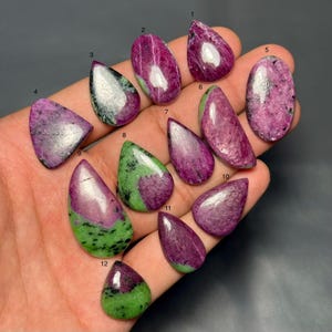 Puede incluir: Una colección de piedras preciosas de rubí zoisita pulidas en varias formas, incluyendo lágrimas y óvalos. Las piedras muestran una mezcla de colores rosa intenso y verde, con algunas mostrando un efecto veteado. Las piedras preciosas se exhiben sobre un fondo oscuro.