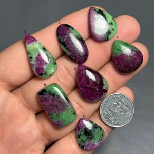 May include: Seven polished ruby zoisite gemstones in different shapes, such as oval, teardrop, and rectangular. The stones show a mix of deep red, green, and black colours. A coin is included for size reference.