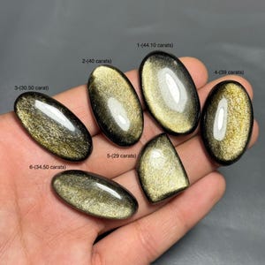 May include: Six polished golden obsidian gemstones of varying shapes and sizes are displayed. Each stone has a unique pattern and is labeled with its carat weight, ranging from 29 to 44.10 carats. The stones are oval, teardrop, and rounded shapes.