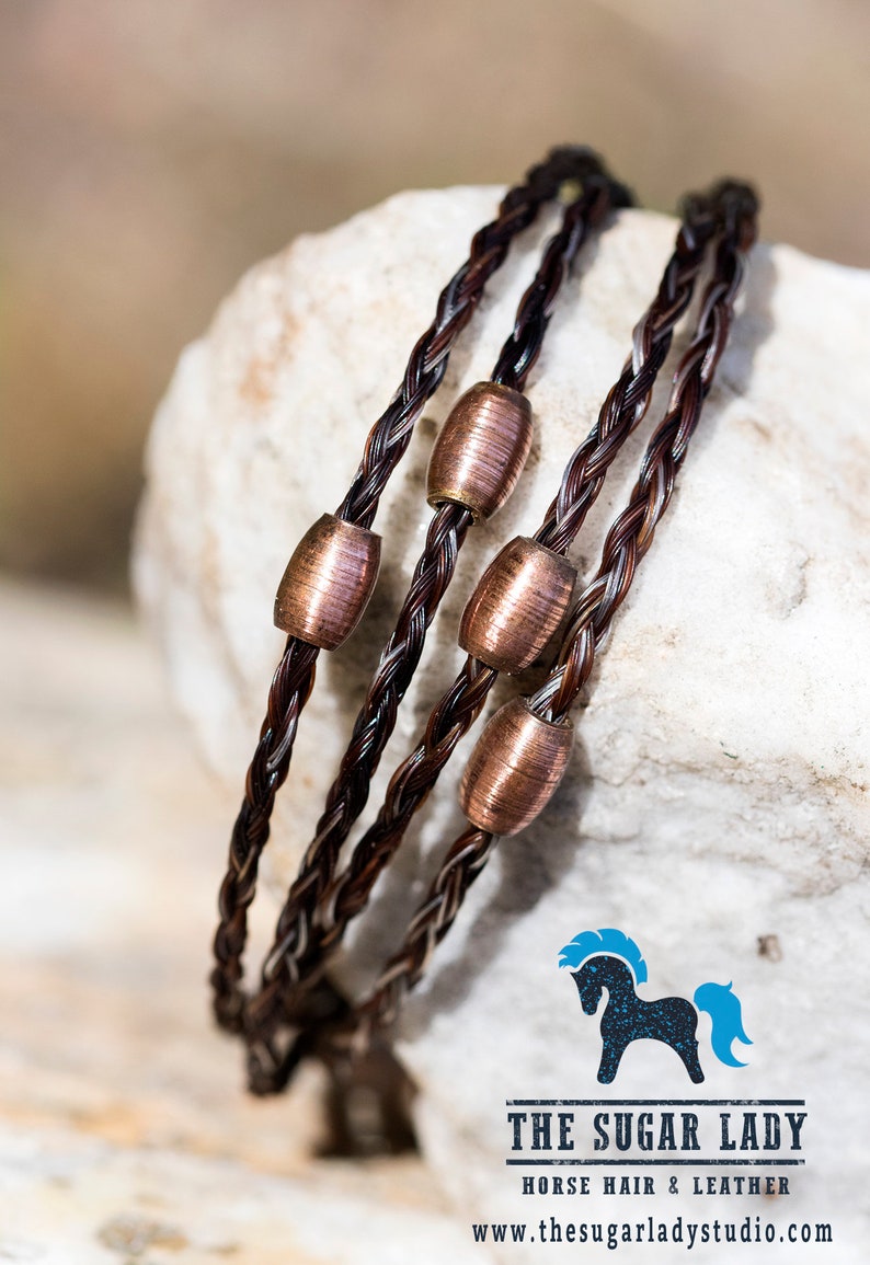 May include: A triple strand bracelet made of brown horsehair with copper beads. The bracelet is on a white rock surface. The text "THE SUGAR LADY HORSE HAIR & LEATHER www.thesugarladystudio.com" is at the bottom of the image.