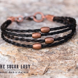 May include: A black horsehair bracelet with copper beads. The bracelet is braided and has a simple clasp. The bracelet is on a brown, textured surface. The text "THE SUGAR LADY HORSE HAIR & LEATHER" is visible below the bracelet.