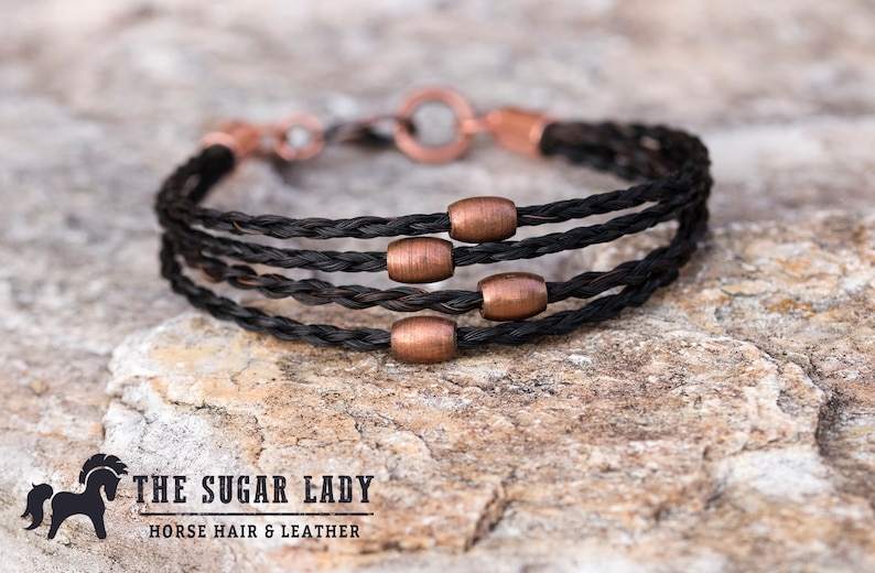 May include: A black horsehair bracelet with copper beads. The bracelet is braided and has a simple clasp. The bracelet is on a brown, textured surface. The text "THE SUGAR LADY HORSE HAIR & LEATHER" is visible below the bracelet.