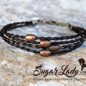 May include: A multi-strand bracelet made of braided horsehair with copper beads. The bracelet is on a brown, textured surface. "Sugar Lady Studio" is written in cursive below the bracelet.