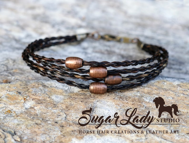 May include: A multi-strand bracelet made of braided horsehair with copper beads. The bracelet is on a brown, textured surface. "Sugar Lady Studio" is written in cursive below the bracelet.