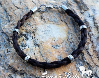 May include: A brown braided horsehair bracelet with silver metal accents. The bracelet has a clasp closure and four decorative rings.