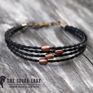 May include: A black braided bracelet with copper beads. The bracelet is made of horsehair and leather. The text "THE SUGAR LADY HORSE HAIR & LEATHER" is visible below the bracelet.