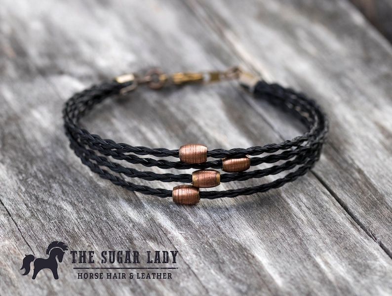 May include: A black braided bracelet with copper beads. The bracelet is made of horsehair and leather. The text "THE SUGAR LADY HORSE HAIR & LEATHER" is visible below the bracelet.