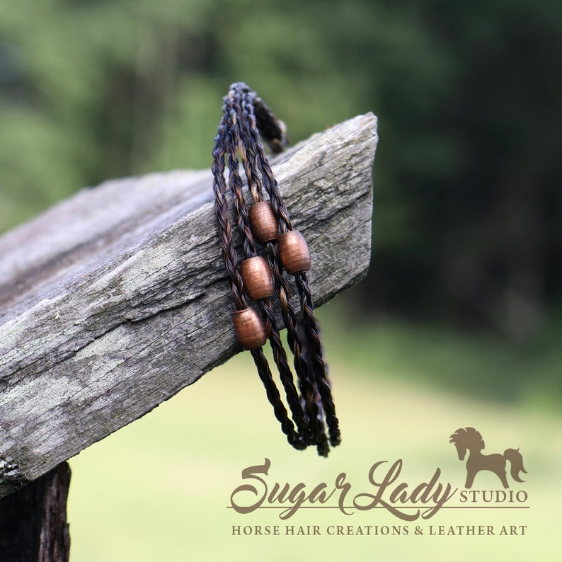 May include: A multi-strand bracelet made of dark brown horsehair with copper beads. The bracelet is draped over a piece of weathered wood. The text "Sugar Lady Studio" is visible in the lower right corner of the image.  Horse Hair Creations & Leather Art.