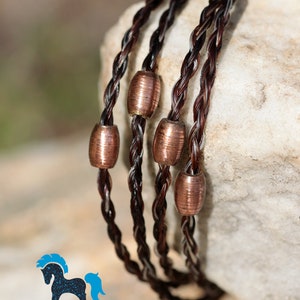 May include: A braided horsehair bracelet with copper beads. The bracelet is brown and white and is wrapped around a light brown rock.