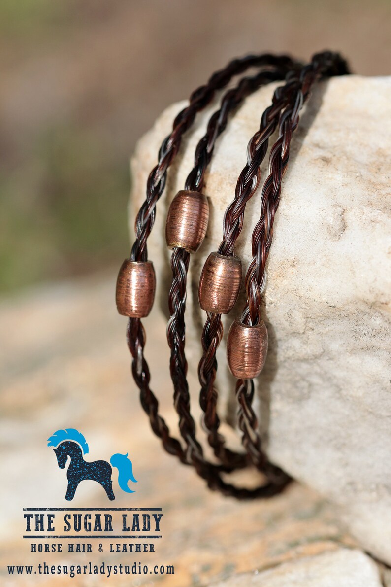 May include: A braided horsehair bracelet with copper beads. The bracelet is brown and white and is wrapped around a light brown rock.