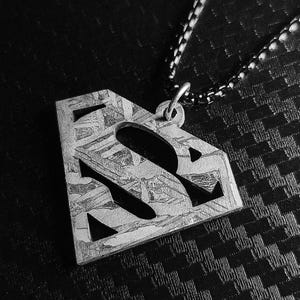 Puede incluir: Un colgante de metal plateado con forma de escudo bordeado de diamantes y una letra S recortada en el centro. La superficie del colgante presenta un patrón grabado cristalino distintivo. Está unido a un collar de cadena de eslabones oscuros y texturizados.