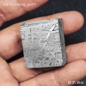 May include: A square-shaped, metallic meteorite with a unique, textured surface. The meteorite is a dark grey colour and weighs 87.6g. The text "METEORITE GIFT" is visible at the top.