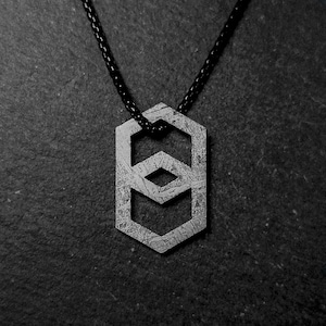 Puede incluir: Un collar con colgante geométrico con un acabado metálico texturizado. El colgante presenta una forma hexagonal con un diseño abierto, que incorpora un patrón de diamantes y galones. El collar tiene un cordón negro. El fondo es una superficie oscura y texturizada.