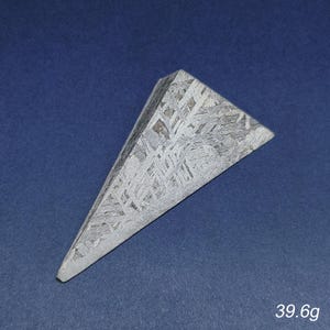 May include: A triangular, metallic meteorite specimen with a patterned surface. The gray-toned stone is shaped like a pyramid, with a flat base and a pointed top. The weight is 39.6 grams.